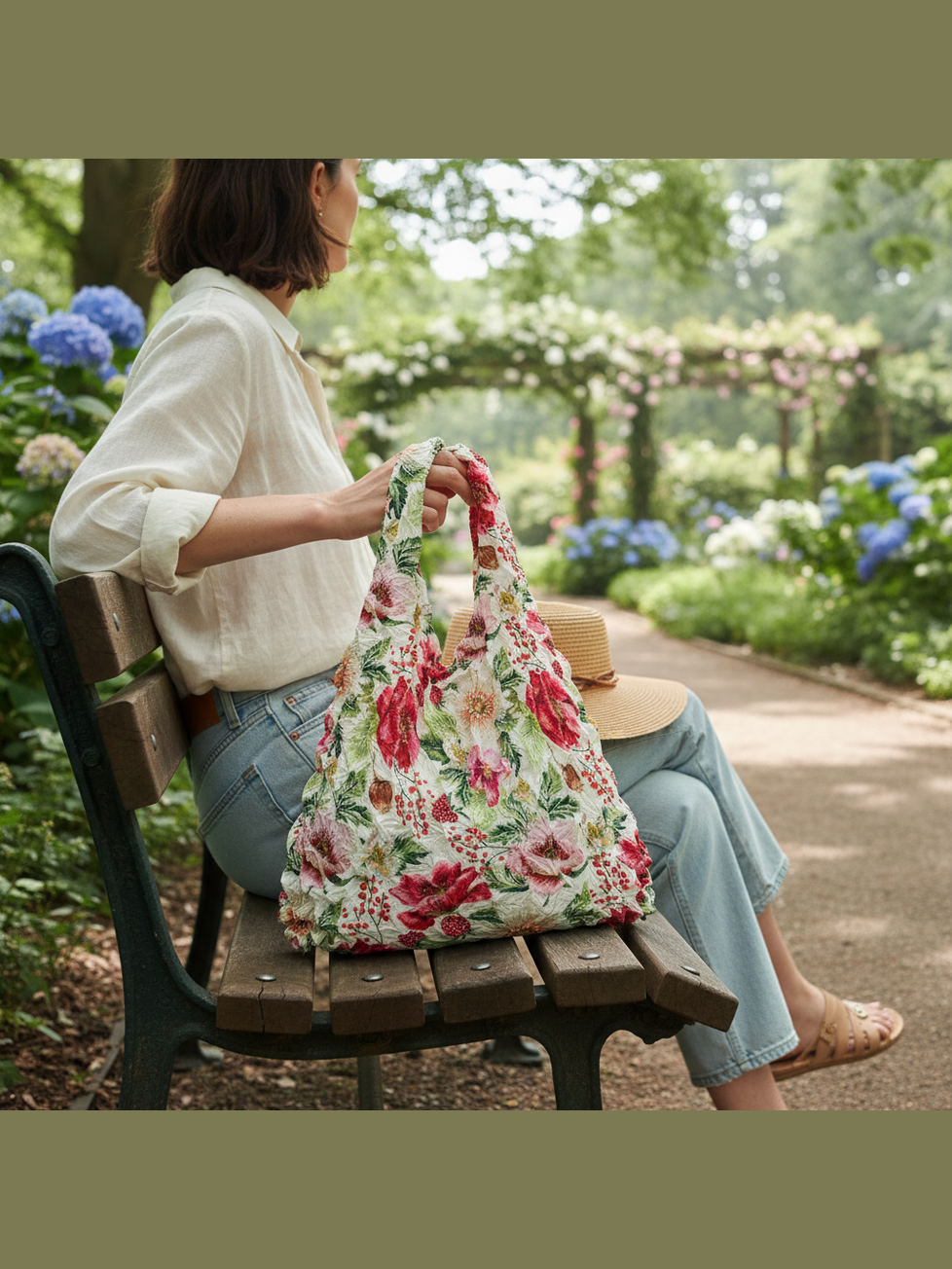 Be Bougie stretchy eco tote bag with a vibrant red and green berries and anemones floral print held on a park bench.