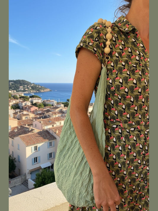 Be Bougie wooden bead bag handle attached to a green fabric tote bag worn by a woman with a coastal town background