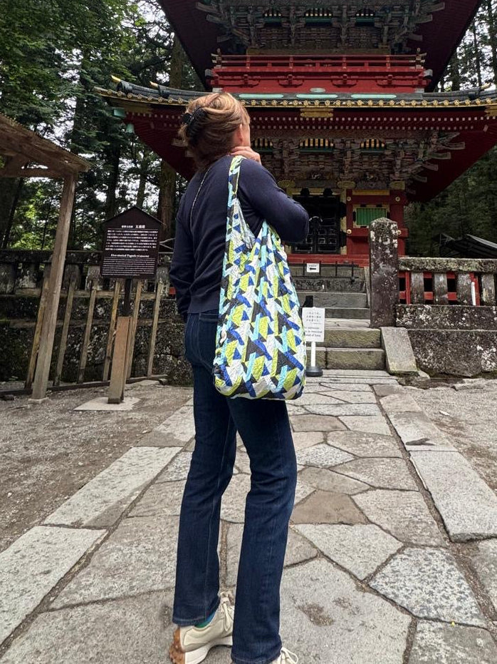 A woman wearing a navy blue long-sleeve top and dark jeans stands on a stone path in a forest, looking up at a large red and green Japanese pagoda while carrying a blue, green, and yellow patterned Maxi tote, by Be Bougie.