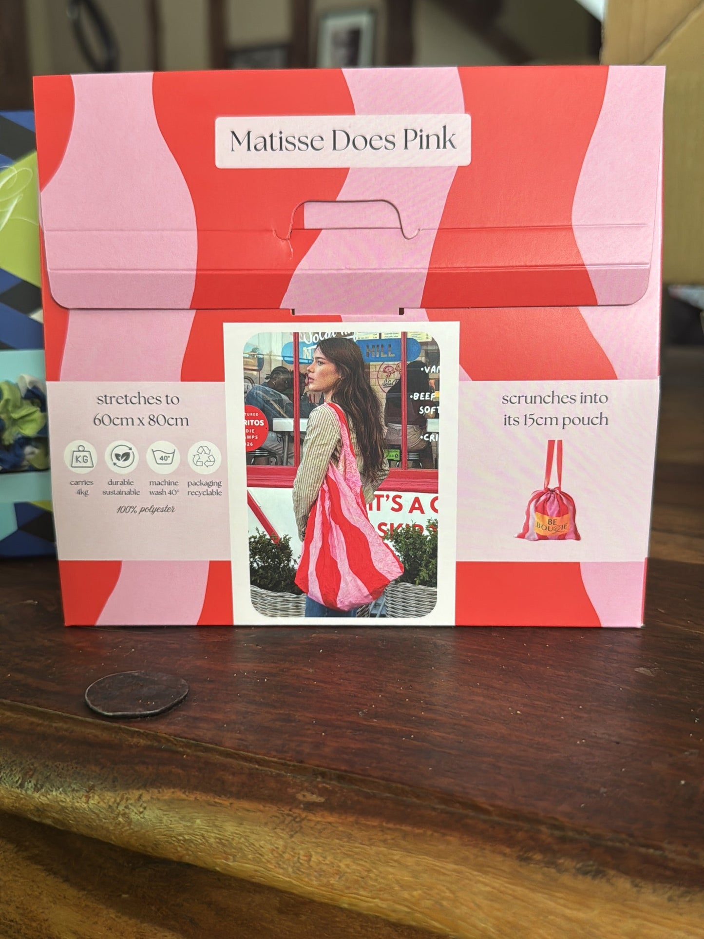 A Matisse Does Pink carded envelope-style pouch for making scarf pouches, displayed on a wooden table. The packaging features a pink and coral wavy pattern with a central photo of a woman wearing a matching pink and red scarf.