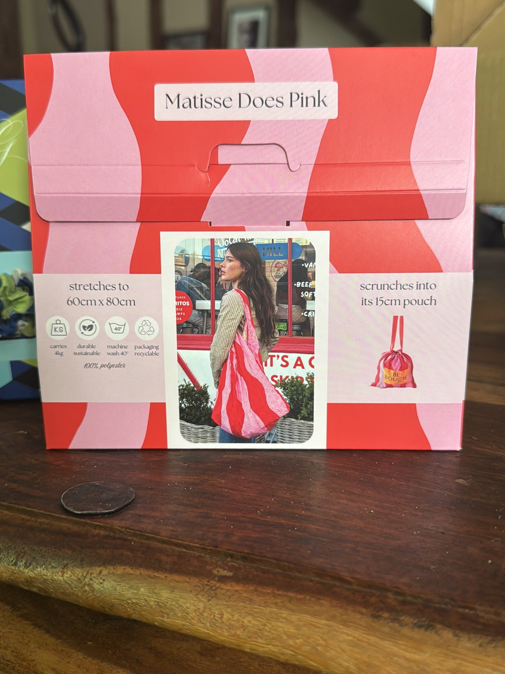 A Matisse Does Pink carded envelope-style pouch for making scarf pouches, displayed on a wooden table. The packaging features a pink and coral wavy pattern with a central photo of a woman wearing a matching pink and red scarf.
