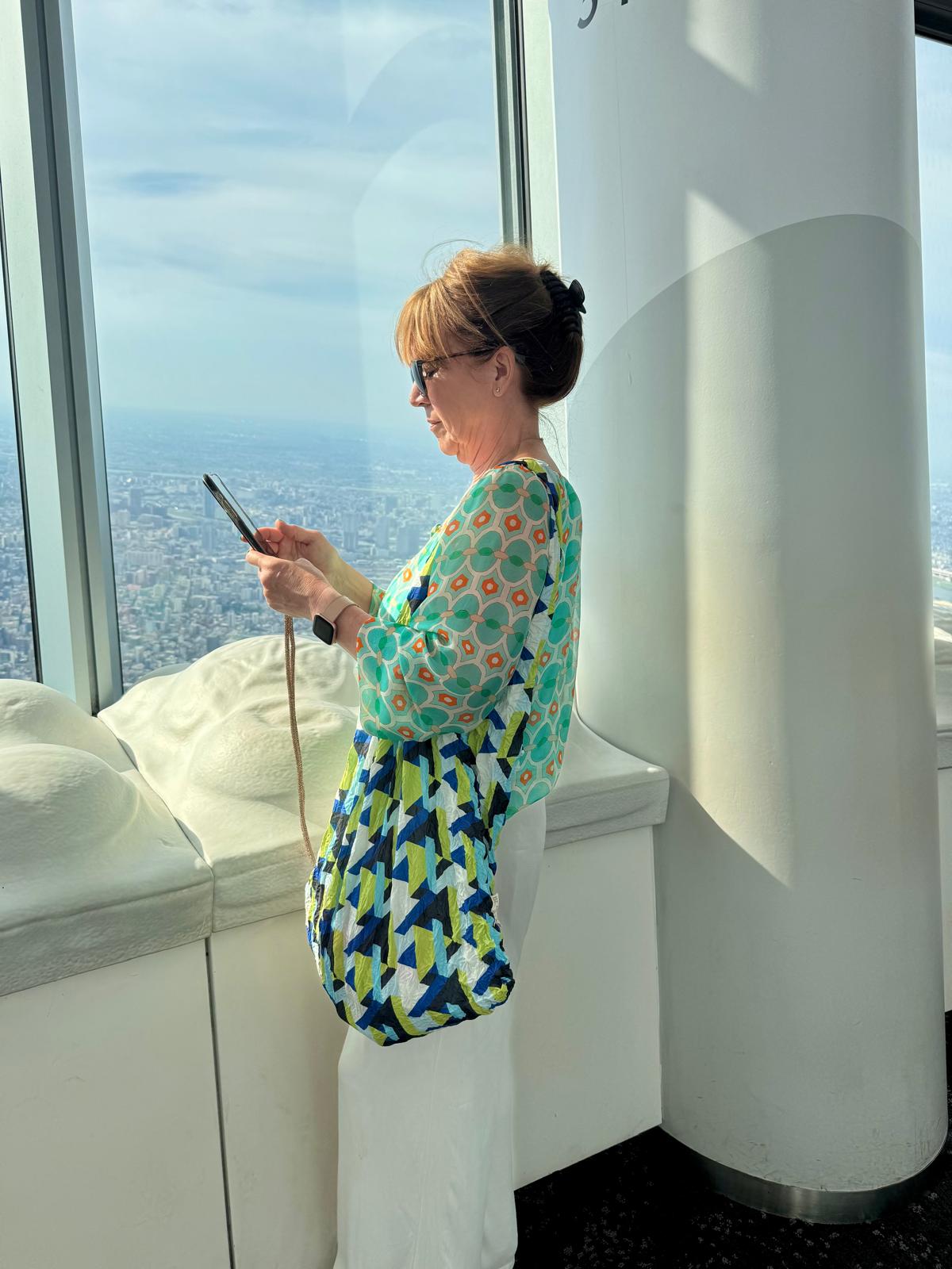Be Bougie gold sparkly cross body phone strap worn by a woman looking at her phone near a city window view