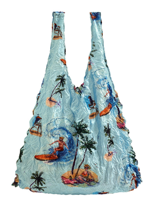 Be Bougie sustainable stretchy tote bag in light blue with tropical beach, surfing, and palm tree illustrations.